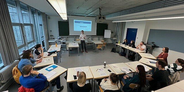 Seminarraum mit einem Vortragenden und Studierenden im Plenum.