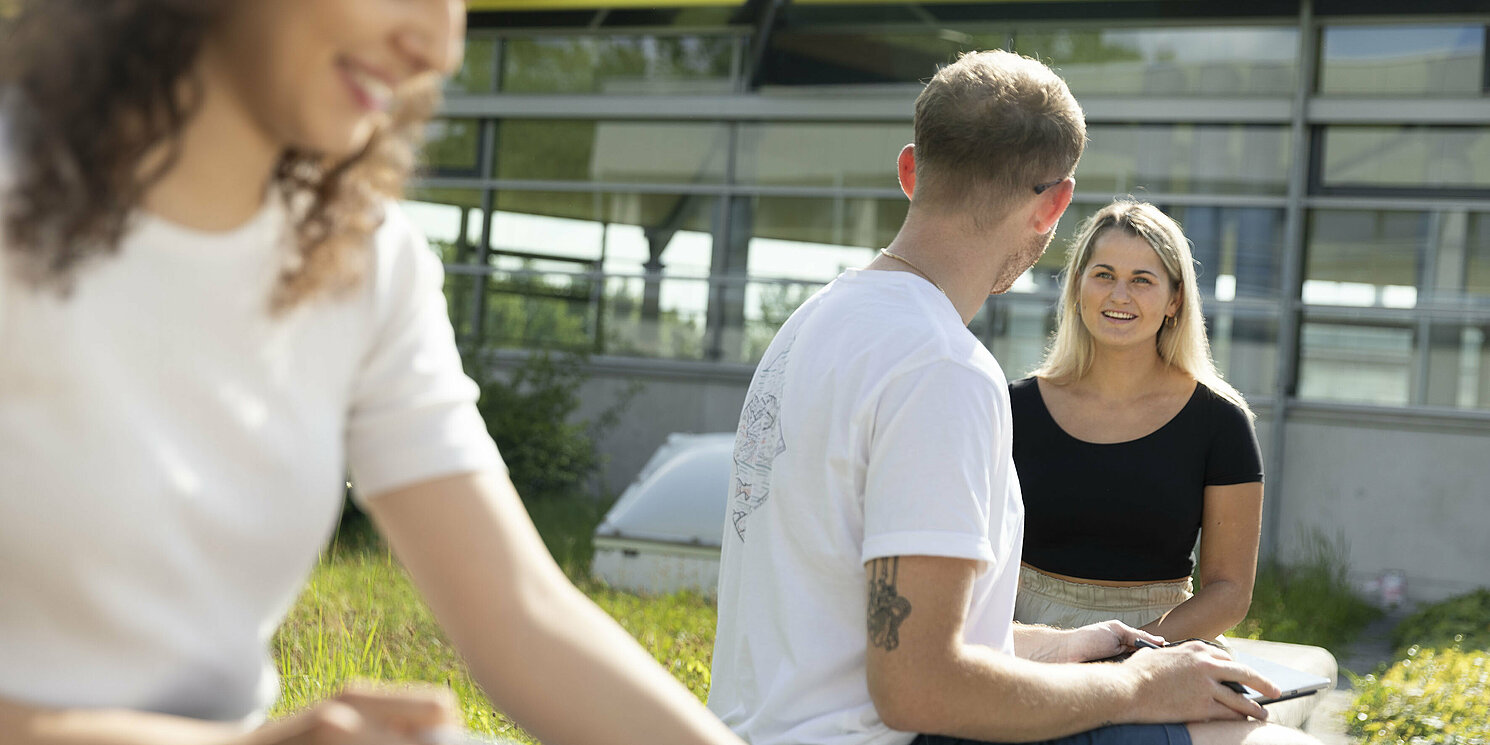 Drei  Personen sitzen im Sommer draußen vor dem OTH Gebäude, zwei Personen unterhalten sich.