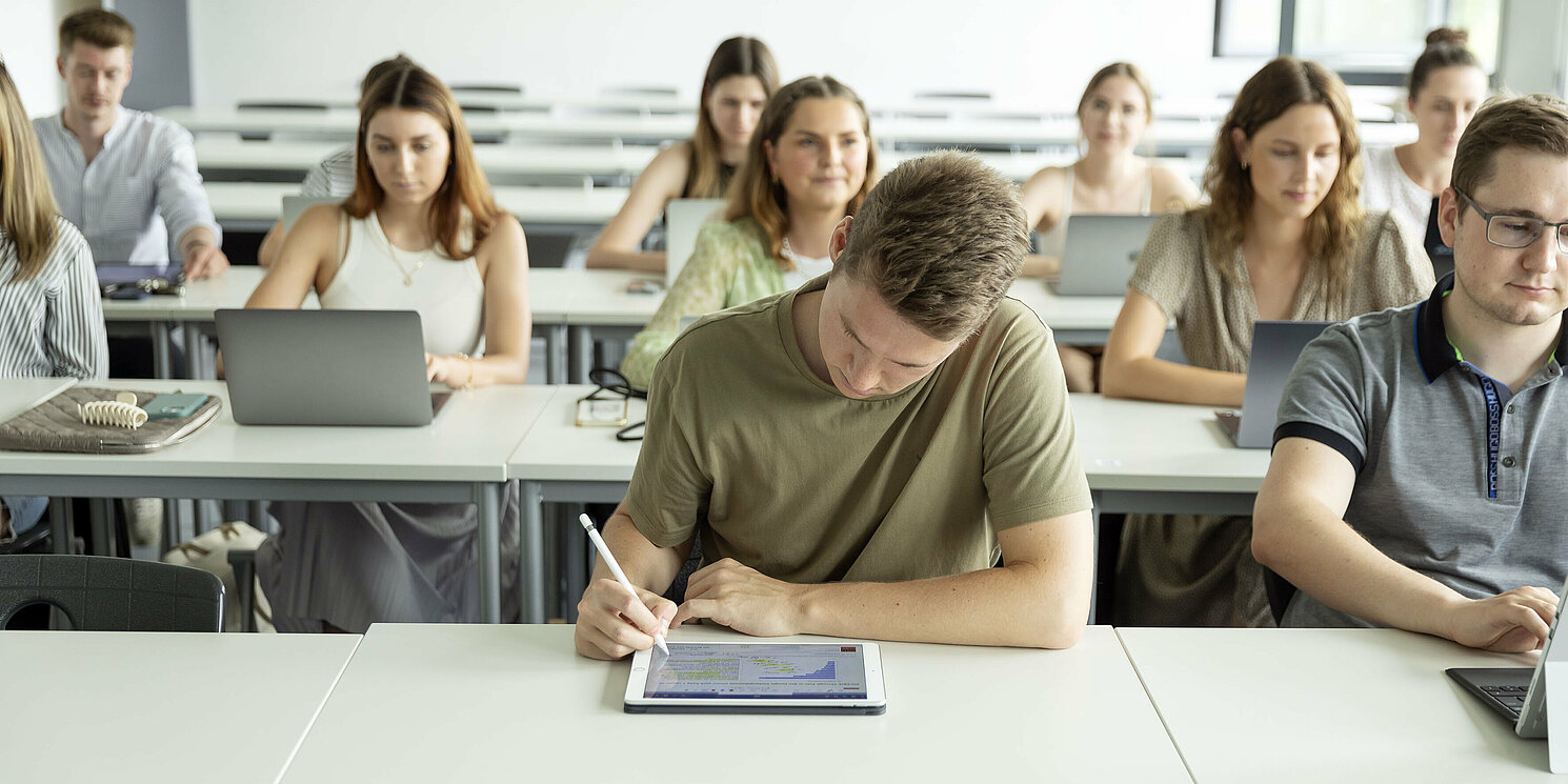 Eine Studierendengruppe in einem Hörsaal. In der ersten Reihe schreibt eine Person auf einem Tablet.