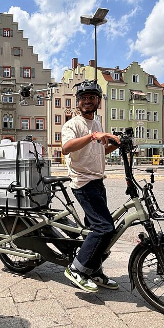 Links im Bild ein Studierender der Die Drohne steuert, Mittig im Bild ein Student auf dem Sensorbike, das auf dem Gepäckträger mit einer großen Box, in der sich der COmputer befindet, aufgestattet ist. Umgebung ist der Arnulfsplatz in Regensburg, eine Innenstadtszene mit bunten Altstadtfassaden im Hintergrund