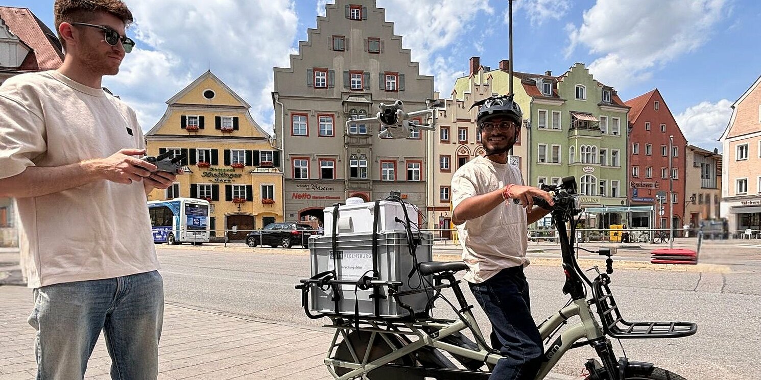 Links im Bild ein Studierender der Die Drohne steuert, Mittig im Bild ein Student auf dem Sensorbike, das auf dem Gepäckträger mit einer großen Box, in der sich der COmputer befindet, aufgestattet ist. Umgebung ist der Arnulfsplatz in Regensburg, eine Innenstadtszene mit bunten Altstadtfassaden im Hintergrund