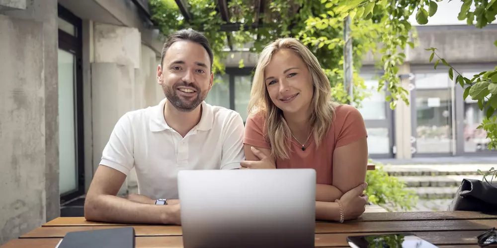 Zwei Studierende sitzen vor einem Laptop und lächeln in die Kamera