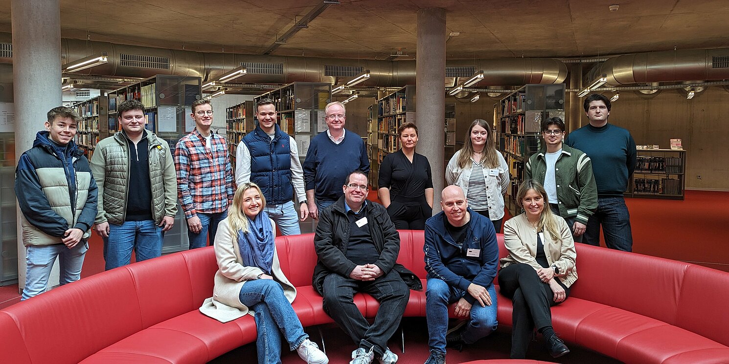 Studierende stehen und sitzen versammelt um die rote Couch in der Hochschul-Bibliothek.