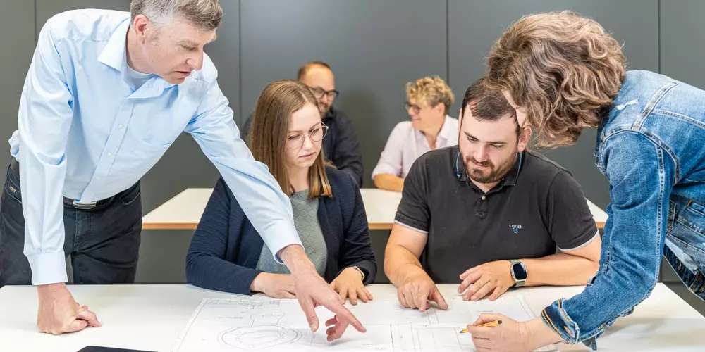 Studierende und ihr Dozent arbeiten an einem Tisch mir verschiedenen Arbeitsunterlagen.