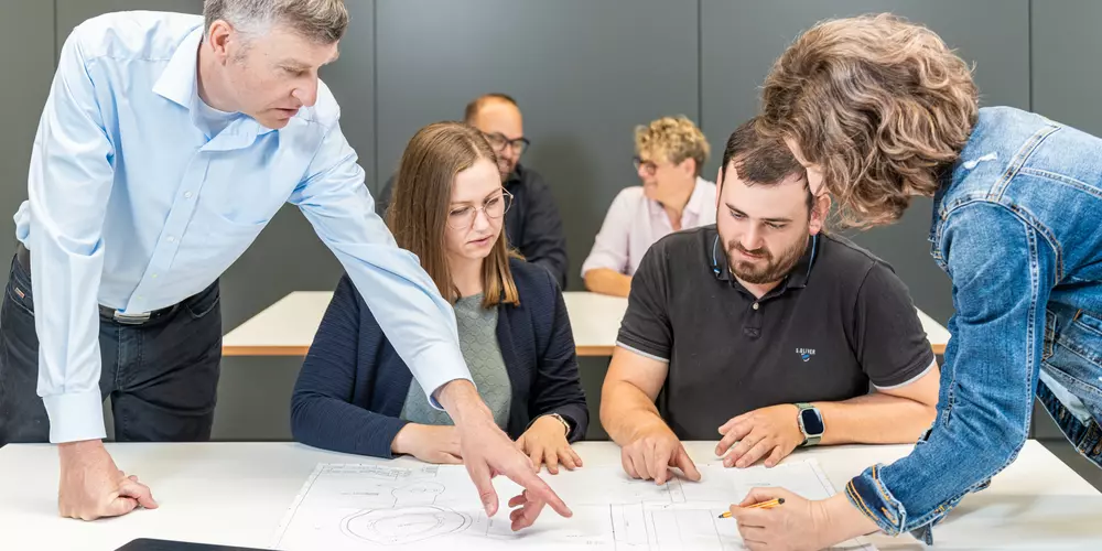 Studierende und ihr Dozent arbeiten an einem Tisch mir verschiedenen Arbeitsunterlagen.