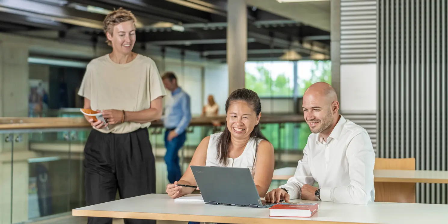 Business Administration – Leadership, Management and Transformation berufsbegleitend studieren Zwei Studierende lernen an einem Tisch und eine dritte Studentin kommt vorbei.