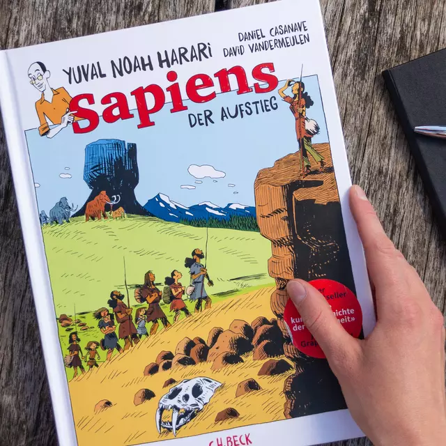 Das Bild zeigt die Grafiknovelle "Sapiens - Der Aufstieg". Sie liegt neben einer Kaffeetasse und einem Notizbuch geschlossen auf einem Holztisch.