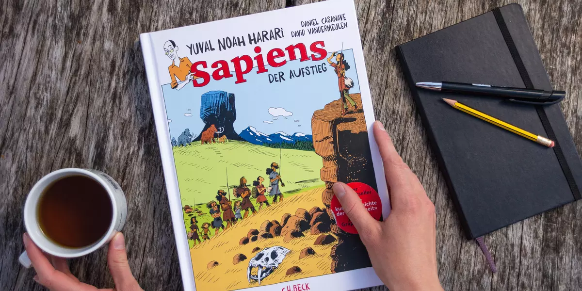 Das Bild zeigt die Grafiknovelle "Sapiens - Der Aufstieg". Sie liegt neben einer Kaffeetasse und einem Notizbuch geschlossen auf einem Holztisch.