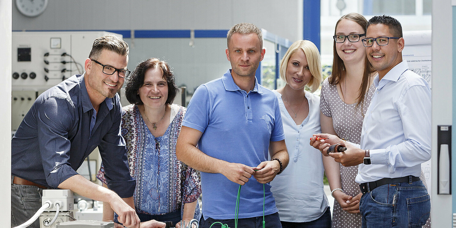 Sechs Personen stehen in einem Labor an technischen Geräten in einer Gruppe beisammen