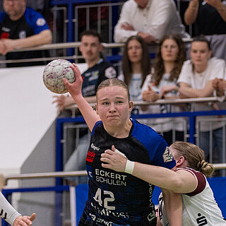 Wettkampfszene beim Handballspiel in der Halle: Milena Stürenburg in der Mitte, in begriff den Ball zu werden, wird umrahmt von zwei gegnerischen Spielerinnen. Im Hintergrund Zuschauer auf der Tribüne.