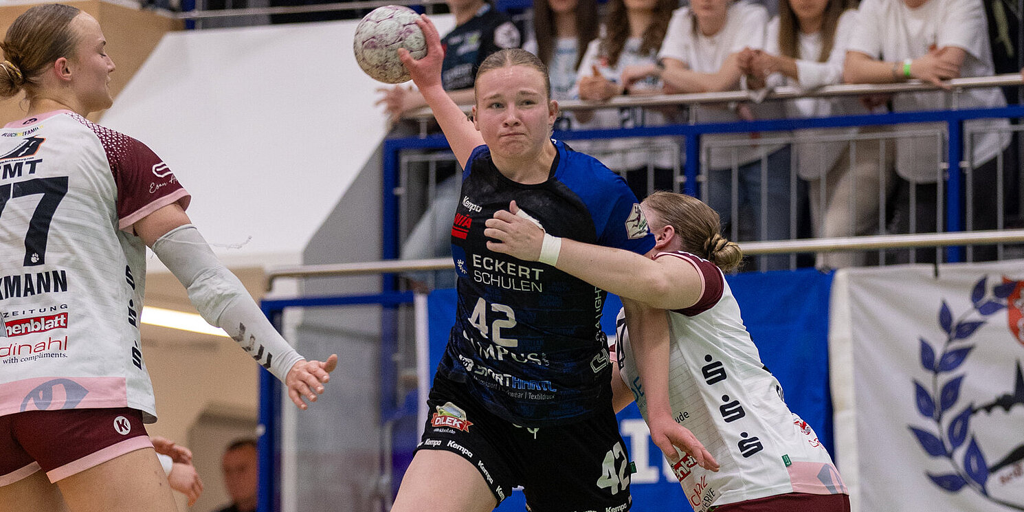 Wettkampfszene beim Handballspiel in der Halle: Milena Stürenburg in der Mitte, in begriff den Ball zu werden, wird umrahmt von zwei gegnerischen Spielerinnen. Im Hintergrund Zuschauer auf der Tribüne.