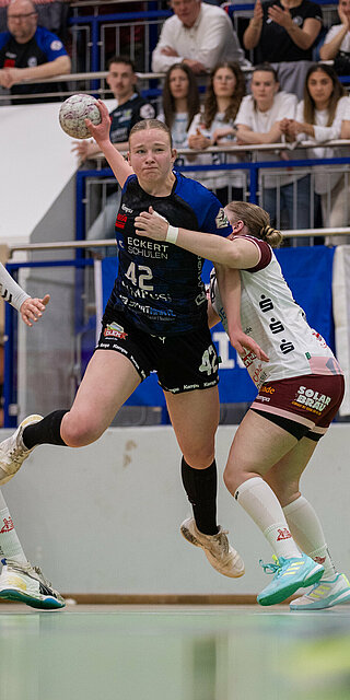 Wettkampfszene beim Handballspiel in der Halle: Milena Stürenburg in der Mitte, in begriff den Ball zu werden, wird umrahmt von zwei gegnerischen Spielerinnen. Im Hintergrund Zuschauer auf der Tribüne.
