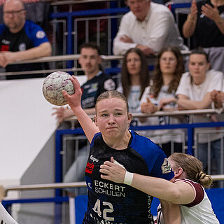 Wettkampfszene beim Handballspiel in der Halle: Milena Stürenburg in der Mitte, in begriff den Ball zu werden, wird umrahmt von zwei gegnerischen Spielerinnen. Im Hintergrund Zuschauer auf der Tribüne.