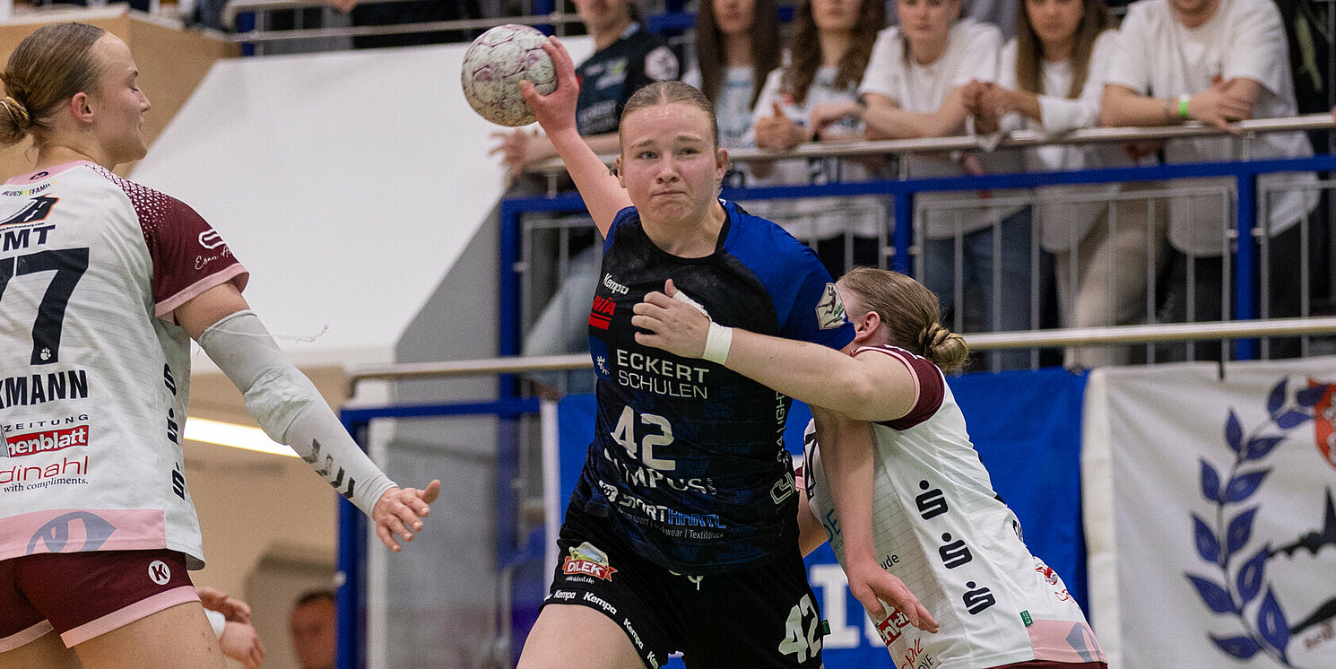 Wettkampfszene beim Handballspiel in der Halle: Milena Stürenburg in der Mitte, in begriff den Ball zu werden, wird umrahmt von zwei gegnerischen Spielerinnen. Im Hintergrund Zuschauer auf der Tribüne.