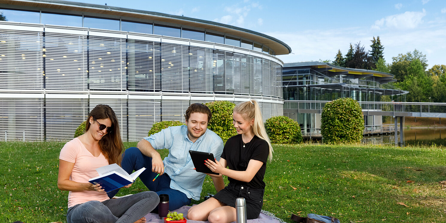 Zentrum für Weitrbildung und Wissensmanagement Drei Personen sitzen auf der Wiese vor der Bibliothek auf einer Decke und lernen gemeinsam