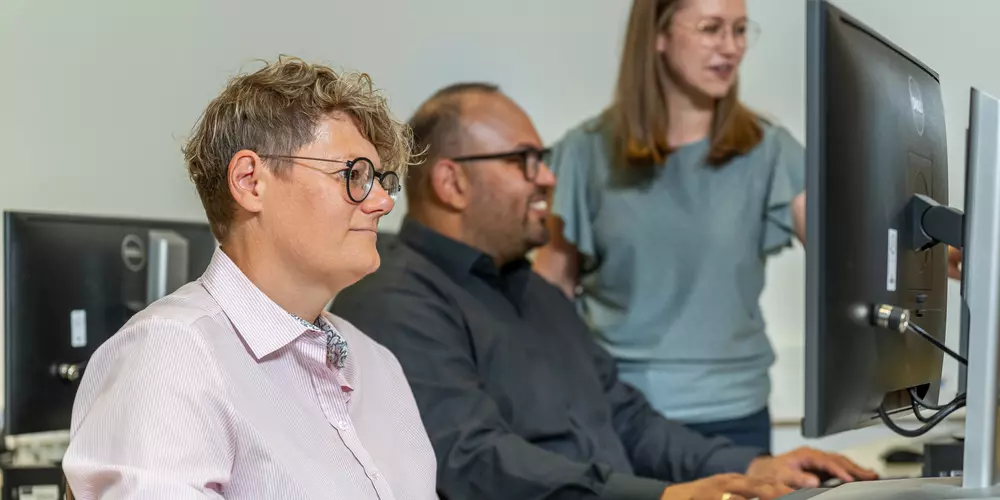 Drei Studierende arbeiten in einem CIP-Pool an ihren Rechnern und unterhalten sich.