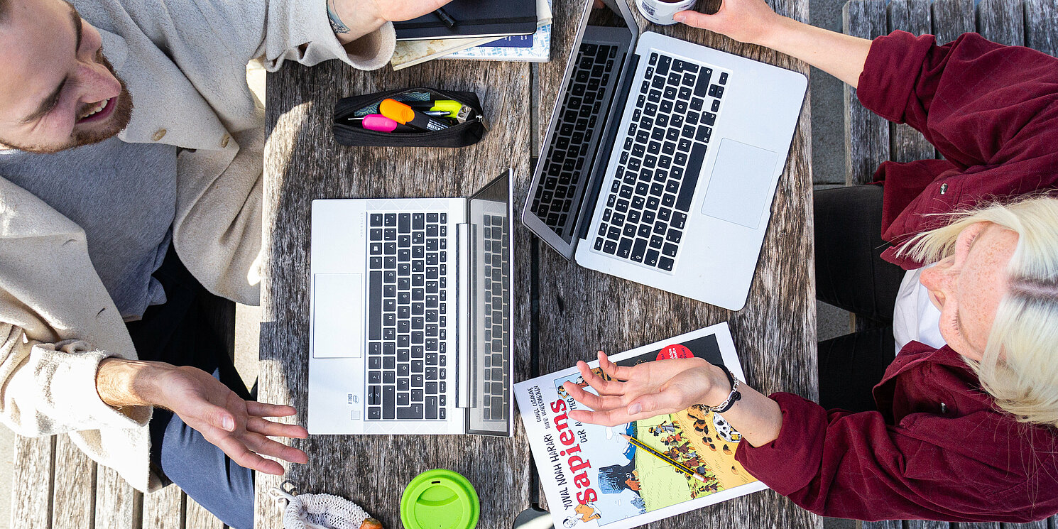 Auf dem Bild sind zwei Menschen zu sehen, die sich gegenübersitzen. Vor ihnen stehen ihre Laptops.