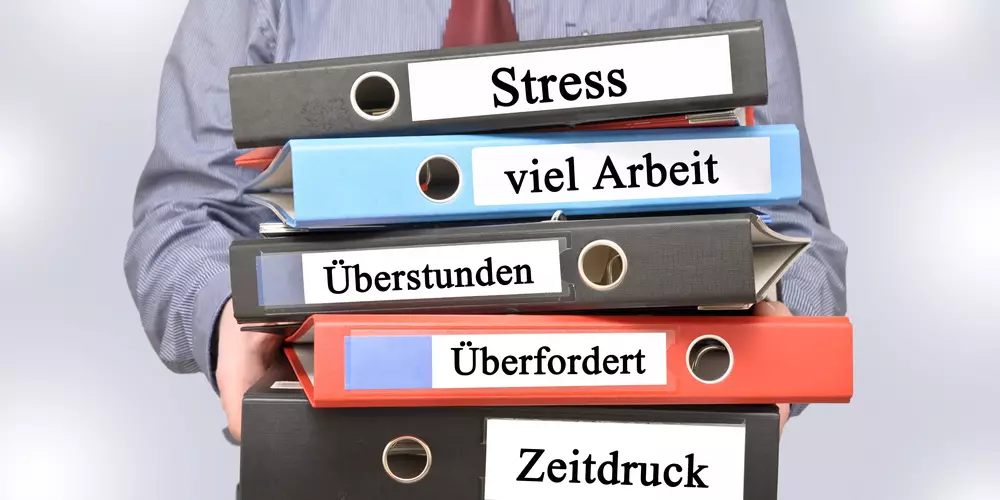 Ein Mann trägt 5 Ordner mit Beschriftungen wie Stress und Überarbeitung.