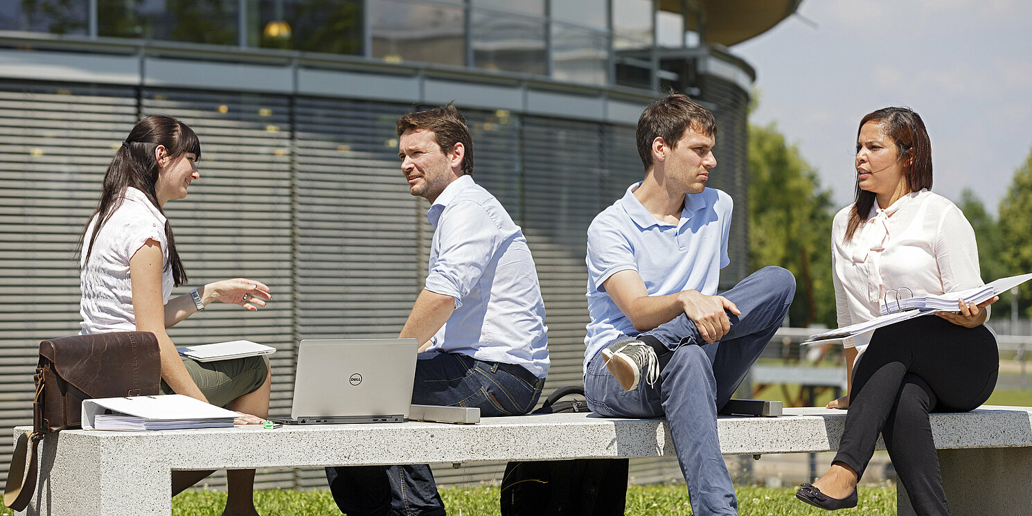 Vier Personen sitzen auf einer Bank vor der Bibliothek über ihren Unterlagen und Laptops und lernen gemeinsam