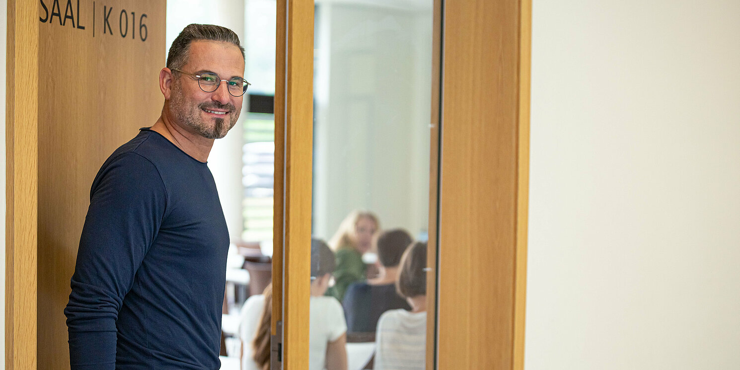 Studentin öffnet Tür zu einem Hörsaal. Darin sitzen bereits Studierende an ihren Plätzen. 