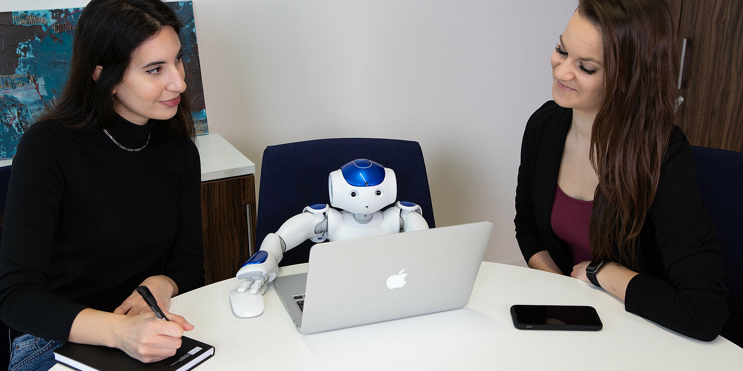 Zwei Personen sitzen mit einem kleinen Roboter an einem Tisch