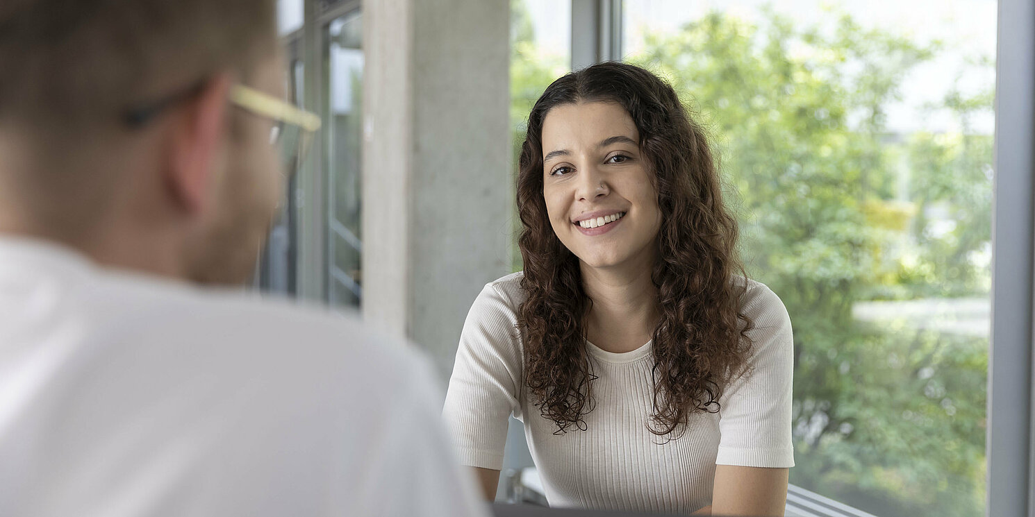 Im Vordergrund sitzt eine dunkelhaarige Frau mit Locken, im Hintergrund sieht man einen Mann mit Brille.