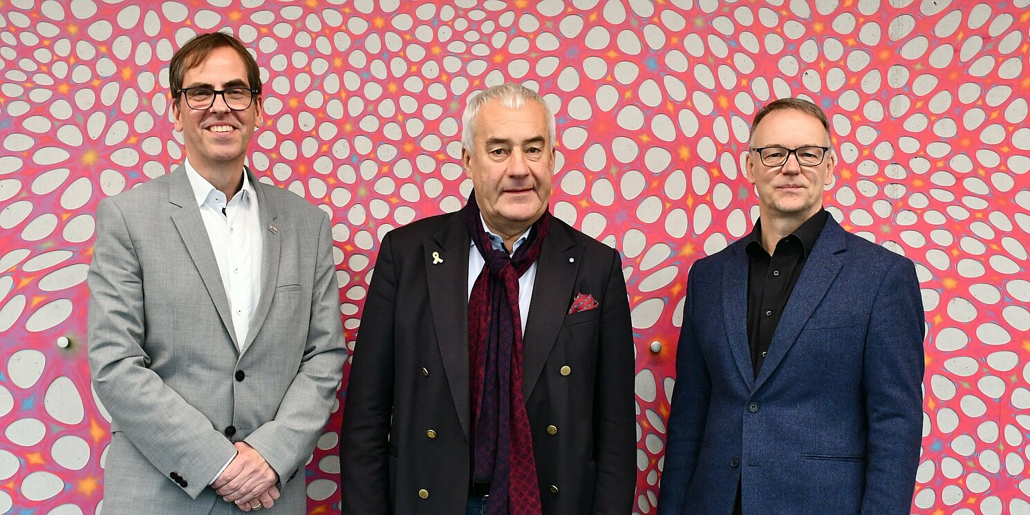Drei Männer stehen vor einer Wand mit buntem Kunstwerk