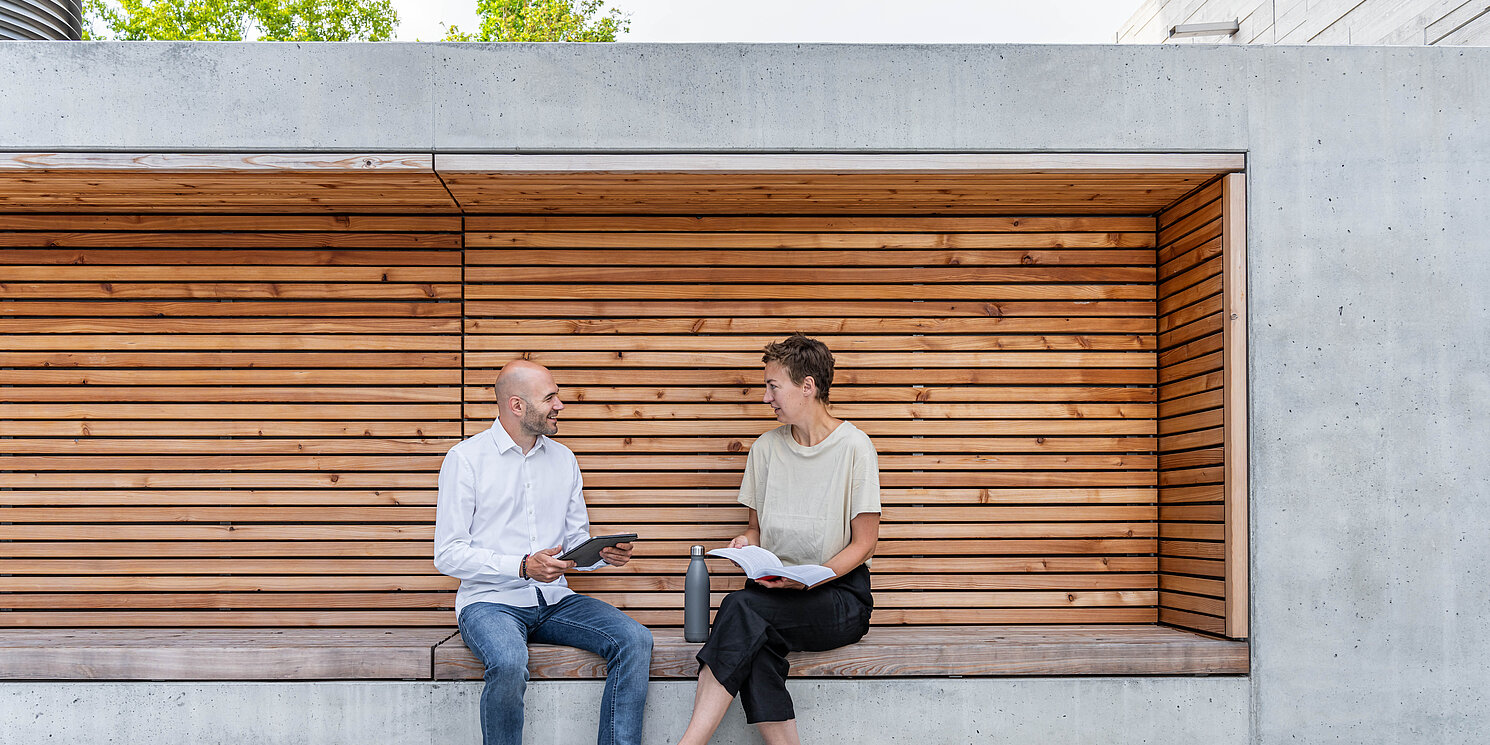 Module aus dem Master Business Administration – General Management berufsbegleitend absolvieren Zwei Studierende sitzen auf einer Bank und unterhalten sich.