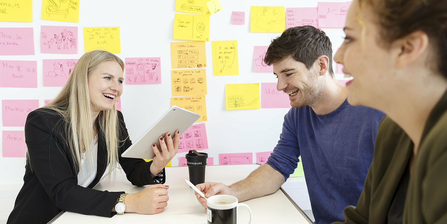 Drei Personen sitzen an einem Tisch und besprechen etwas. Im Hintergrund sind an einer Tafel Post-Its zu sehen. Zwei der Personen sehen auf ein Tablet und ein Smartphone.
