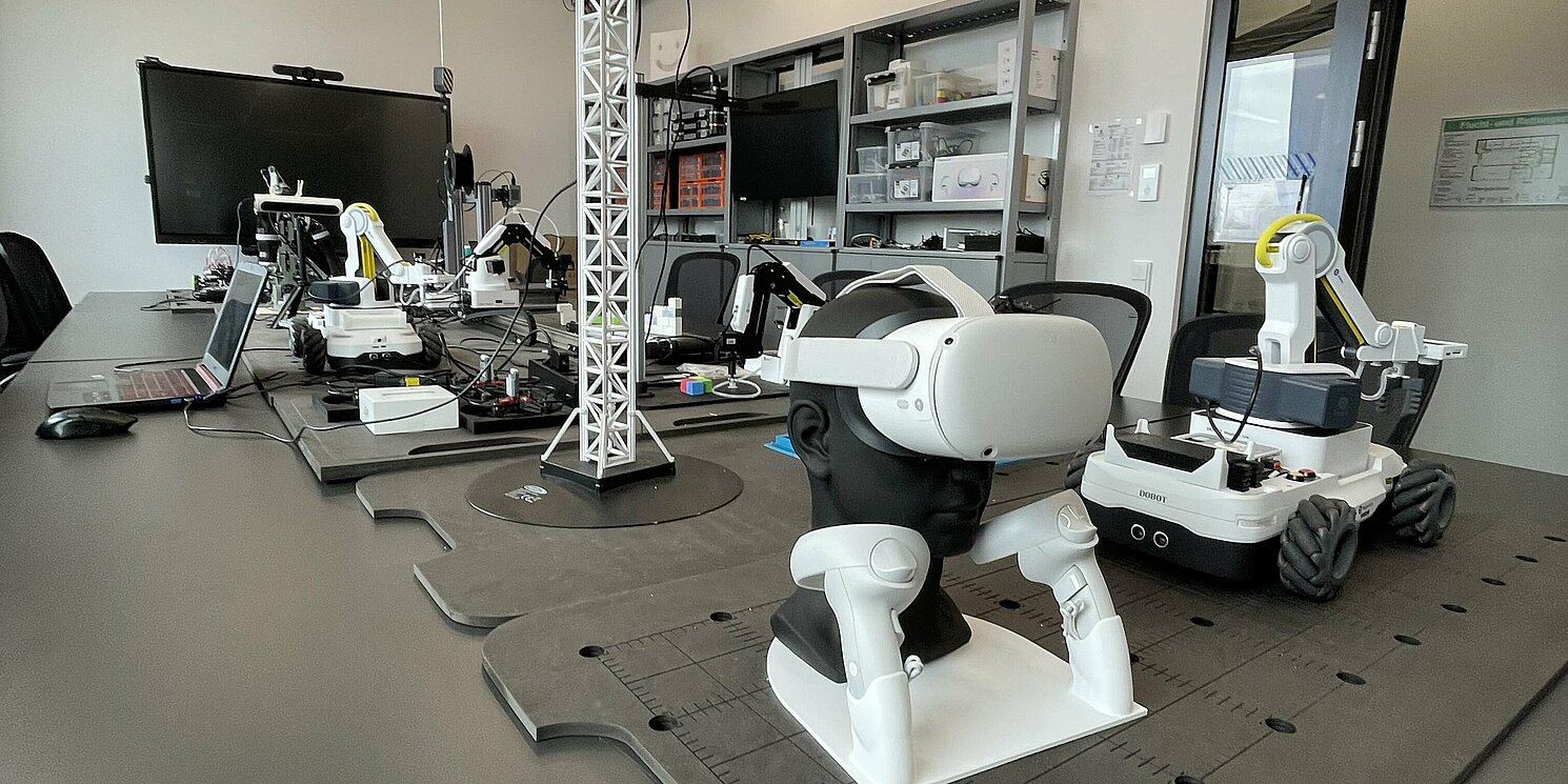 Man sieht ein Labor, in dem verschiedene technische Spielereien aufgebaut sind, u.a. ein Kran oder auch eine VR-Brille mit entsprechenden Joysticks.