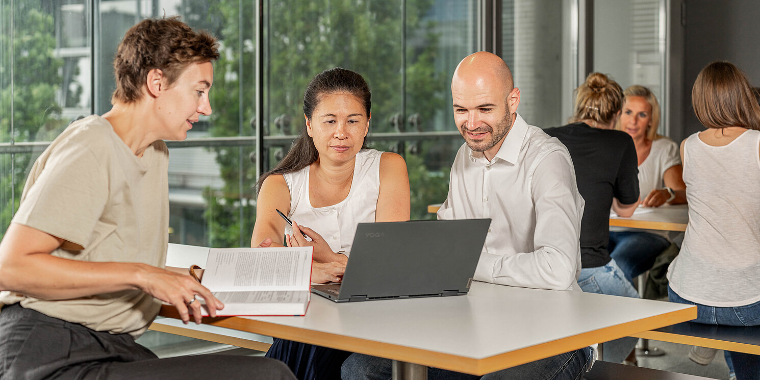 Business Administration – Leadership, Management und Transformation berufsbegleitend studieren Studiereden lernen an Tischen mit ihren Arbeitsmaterialien und unterhalten sich.
