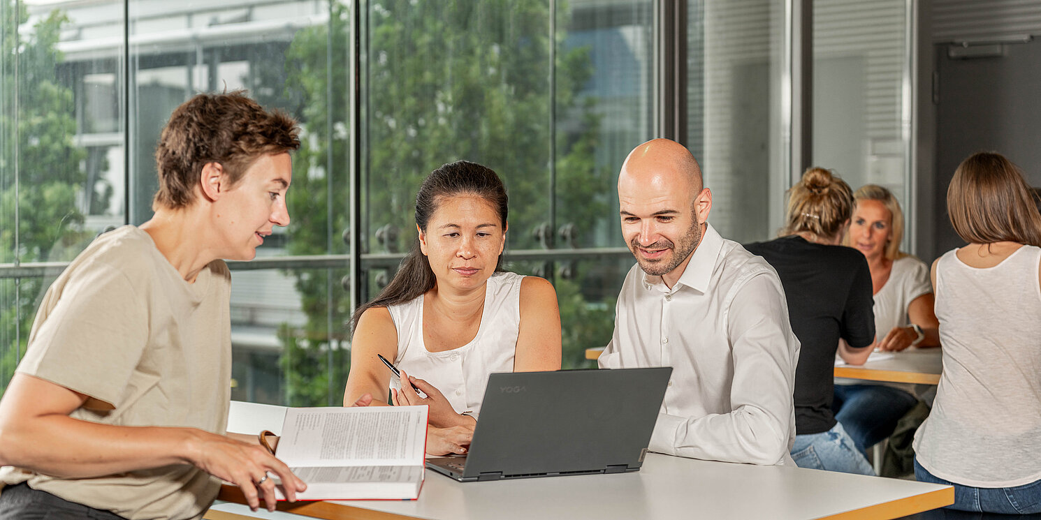 Study Master of Business Administration – Leadership, Management and Transformation part-time Studiereden lernen an Tischen mit ihren Arbeitsmaterialien und unterhalten sich.