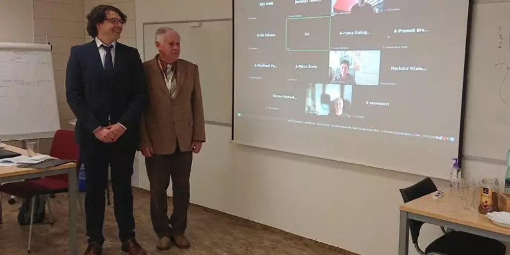Der Doktorand und sein Betreuer stehen vor einer Projektion mit weiteren Teilnehmern der virtuellen Verteidigung