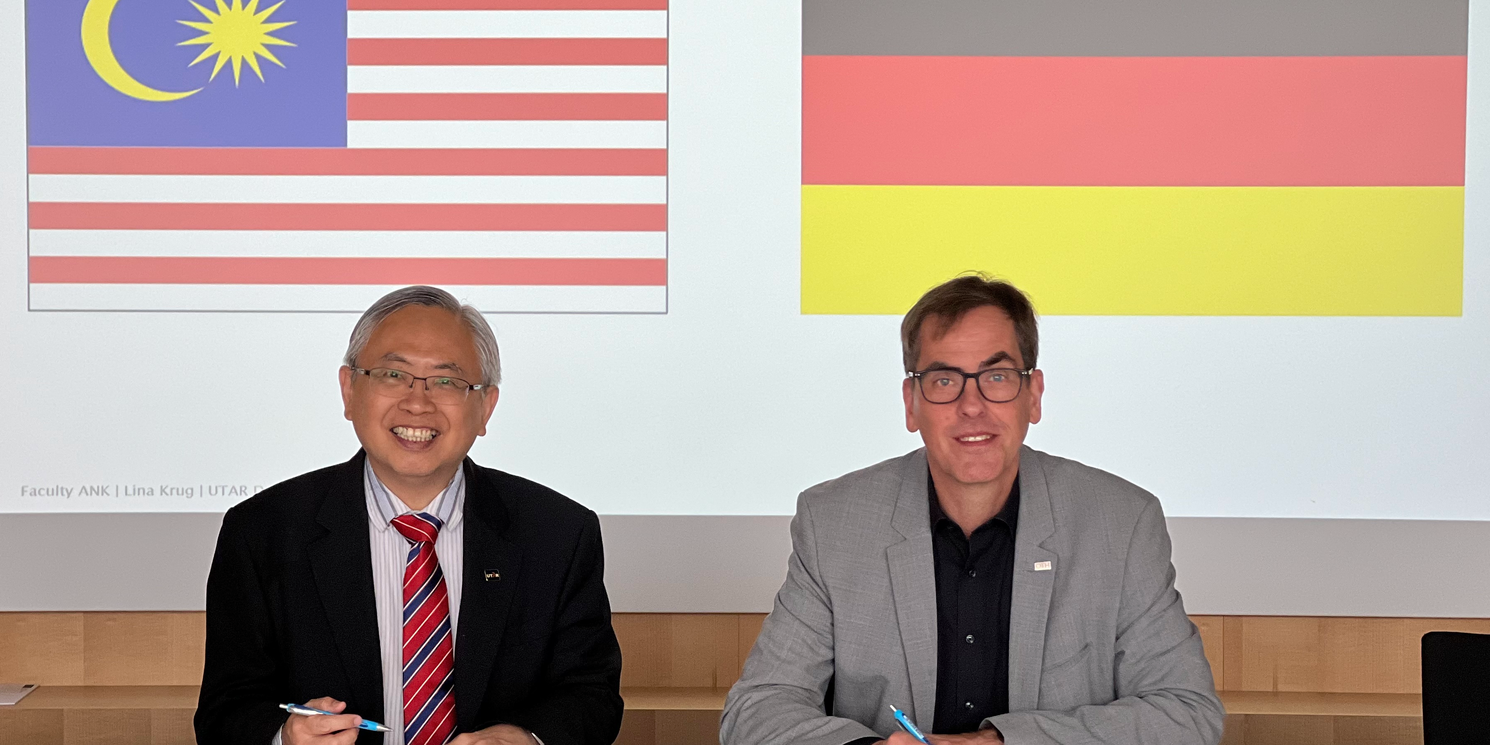 UTAR-Präsident Ir. Professor Dato' Dr. Ewe Hong Tat (l.) und OTH-Präsident Prof. Dr. Ralph Schneider unterzeichnen das Memorandum of Understanding. Sie sitzen nebeneinander vor den Dokumenten und blicken freundlich nach oben in die Kamera. Im Hintergrund sieht man beide Flaggen: Von Malaysia und Deutschland. 