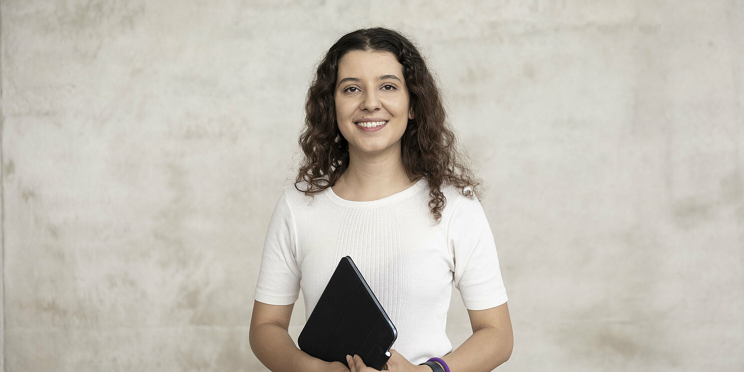 Eine dunkelhaarige Frau mit halblangen Locken. Sie trägt ein weißes Shirt und hält in beiden Händen ein Tablet.