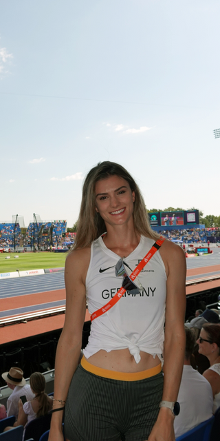 Jana Lakner im Vordergrund mit einem sommerlichen Laufoutfit auf den Besucherrängen des Stadions. Im Hintergrund das Stadium an einem hellen Sommertag. 