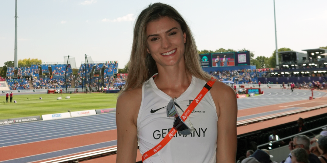 Jana Lakner im Vordergrund mit einem sommerlichen Laufoutfit auf den Besucherrängen des Stadions. Im Hintergrund das Stadium an einem hellen Sommertag. 