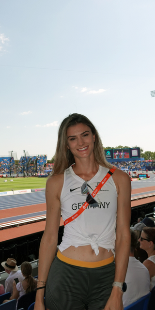 Jana Lakner im Vordergrund mit einem sommerlichen Laufoutfit auf den Besucherrängen des Stadions. Im Hintergrund das Stadium an einem hellen Sommertag. 