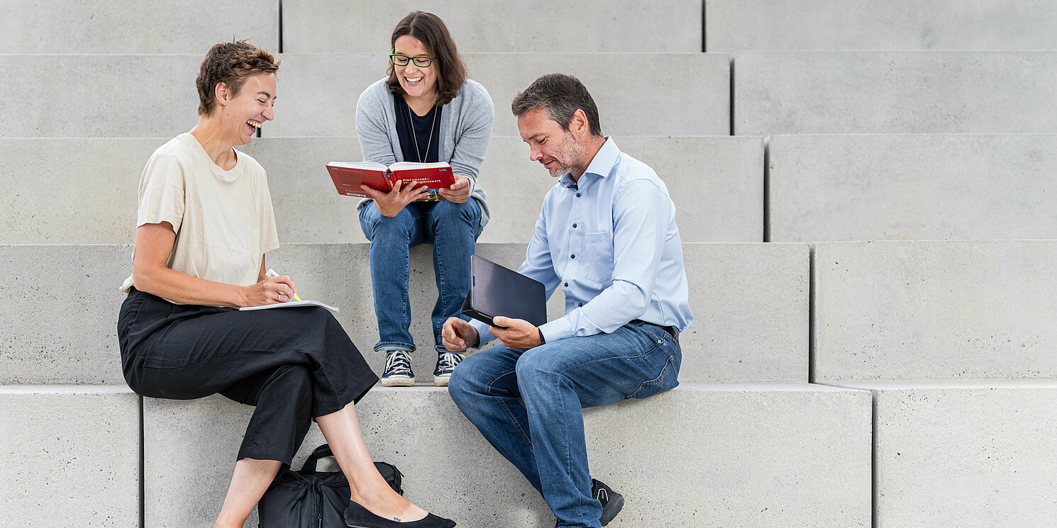 Leitung und Kommunikationsmanagement berufsbegleitend studieren Drei Studierende sitzen mit ihren Lernmaterialien auf einer Treppe.