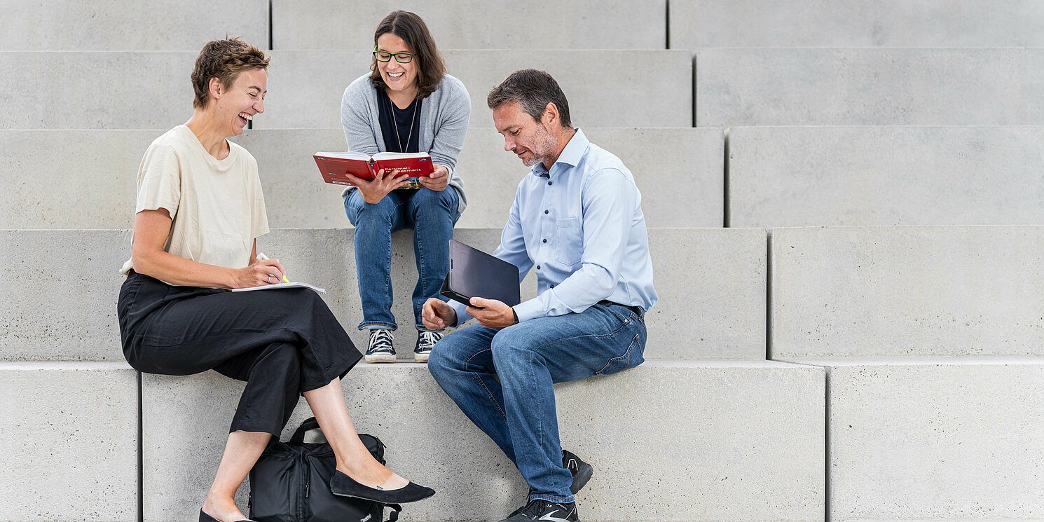 Drei Studierende sitzen mit ihren Lernmaterialien auf einer Treppe. 