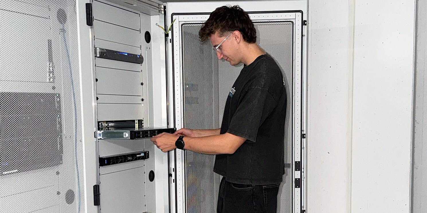 IT-Azubi steht vor einem geöffneten Server und montiert einen Server im Serverschrank. 