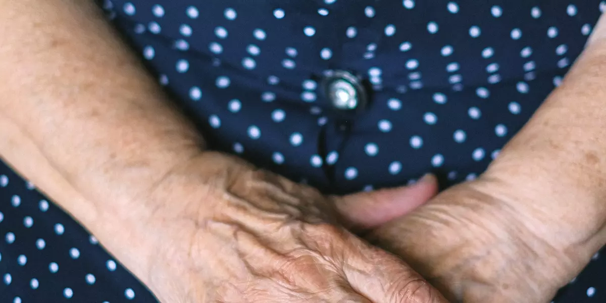 Eine ältere Person mit einem blau gepunkteten Kleid verschränkt die Hände vor dem Körper.