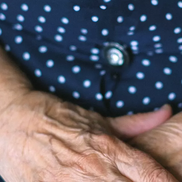 Eine ältere Person mit einem blau gepunkteten Kleid verschränkt die Hände vor dem Körper.