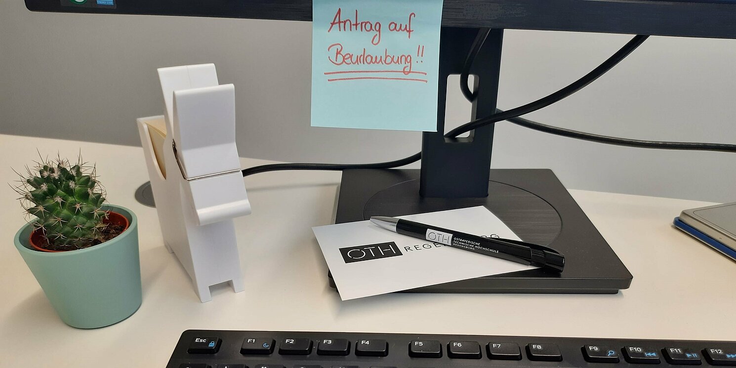 Office desk with a keyboard, pen, decorations and a computer screen with a post-it showing the word „Beurlaubung“ (leave of absence).