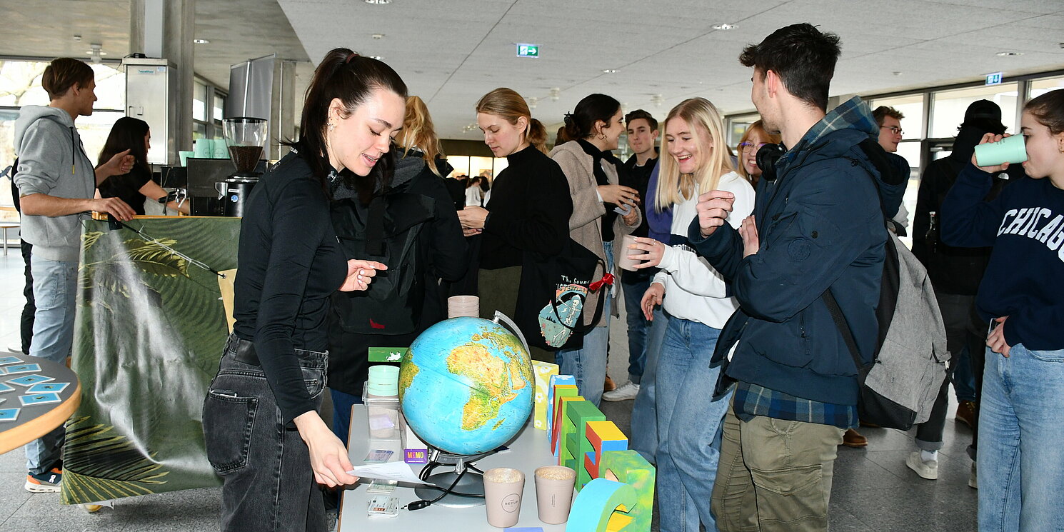 Eine Frau steht an einem Tisch, auf dem ein Globus steht. Davor stehen Menschen, die einen Kaffeebcher in der Hand halten.