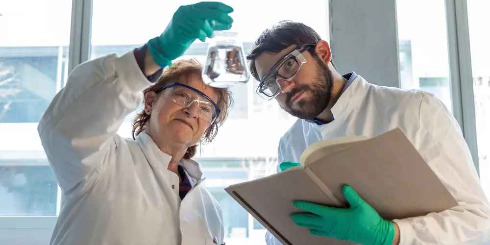 A woman and a man look at an Erlenmeyer flask that the woman is holding up. The man is holding a notebook.