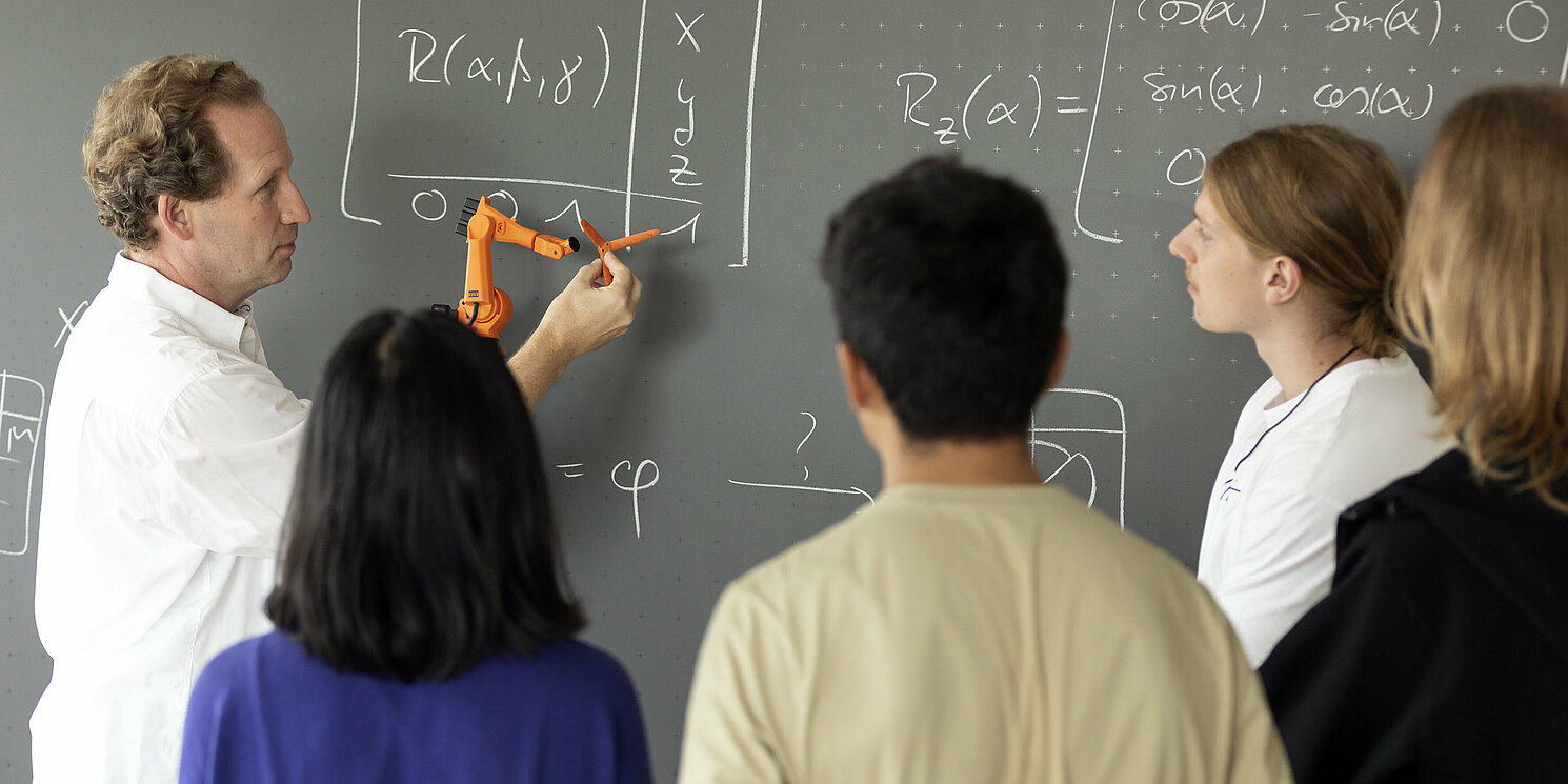 Studierenden wird etwas mit einem kleinen Roboter an einer Tafel erklärt