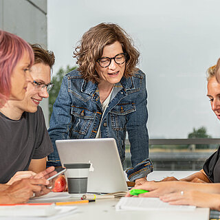 Eine Gruppe von vier Studierenden lernt sitzt zusammen mit zahlreichen Unterlagen an einem Tisch. Angeleitet werden Sie von einer älteren Person, evtl. einer Dozentin, die mit ihrem Laptop an der Stirnseite des Tisches steht. Sie scheinen ein Thema zu diskutieren. 