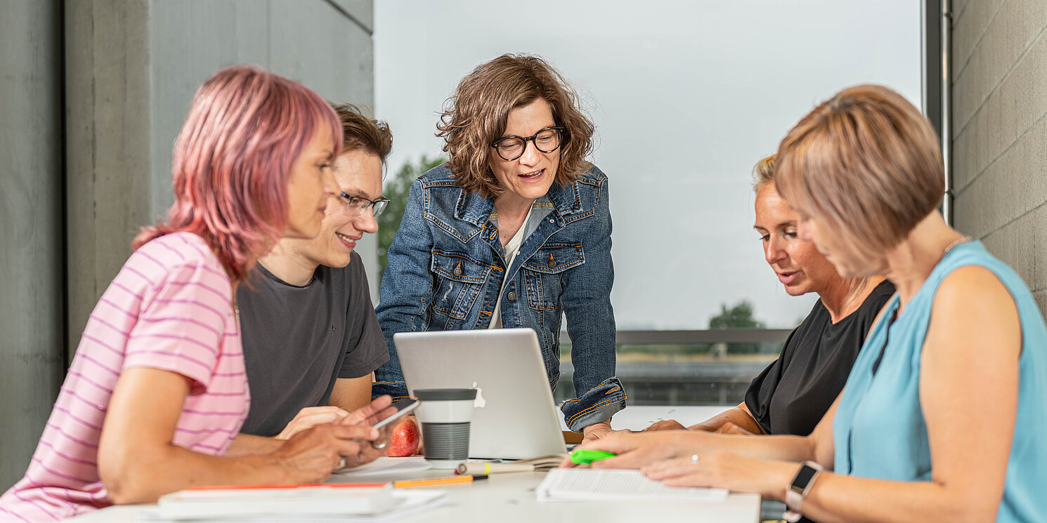 Eine Gruppe von vier Studierenden lernt sitzt zusammen mit zahlreichen Unterlagen an einem Tisch. Angeleitet werden Sie von einer älteren Person, evtl. einer Dozentin, die mit ihrem Laptop an der Stirnseite des Tisches steht. Sie scheinen ein Thema zu diskutieren. 