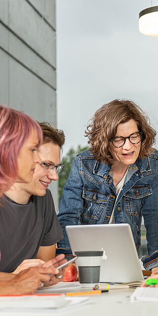 Eine Gruppe von vier Studierenden lernt sitzt zusammen mit zahlreichen Unterlagen an einem Tisch. Angeleitet werden Sie von einer älteren Person, evtl. einer Dozentin, die mit ihrem Laptop an der Stirnseite des Tisches steht. Sie scheinen ein Thema zu diskutieren. 